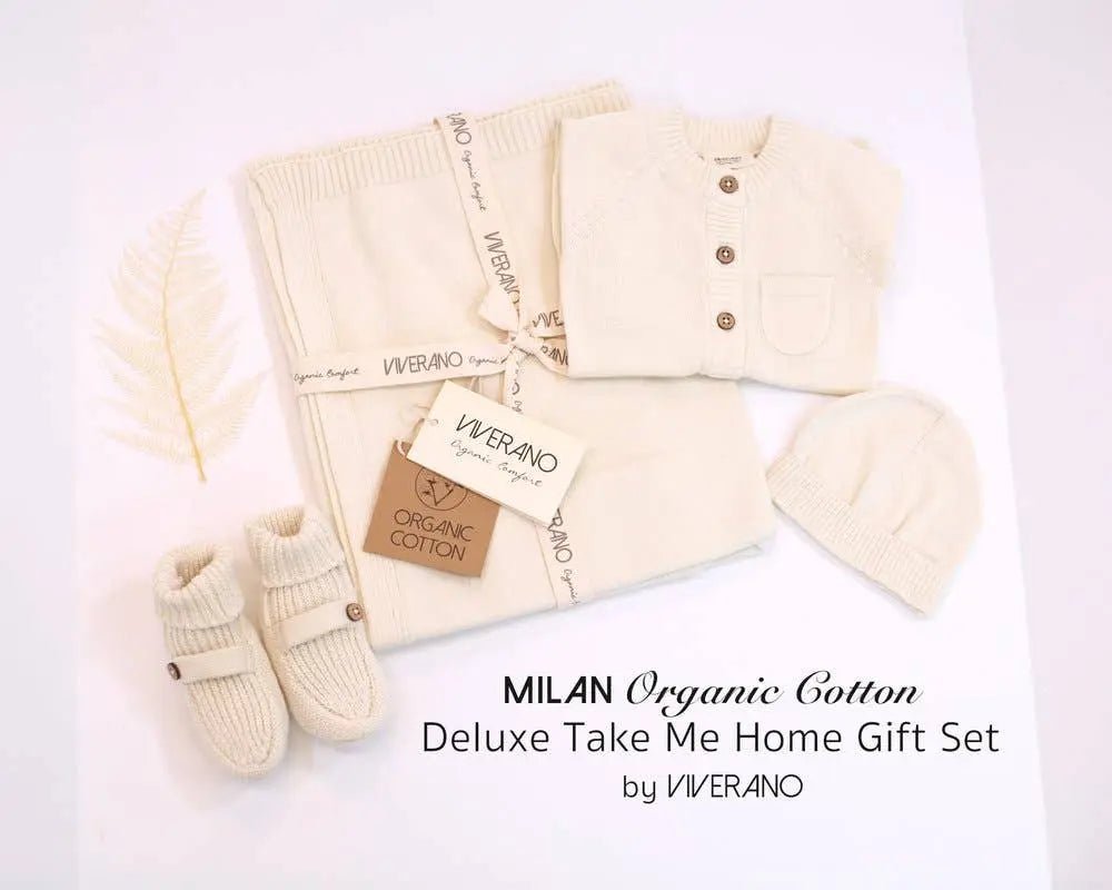 This image displays the "Milan Organic Cotton Deluxe Take Me Home Gift Set" by Viverano. The set includes a soft, cream-colored knit baby cardigan with wooden buttons, matching pants, a beanie, and booties, all made from organic cotton. The items are arranged neatly with branded packaging, emphasizing the luxurious and eco-friendly nature of the gift set.