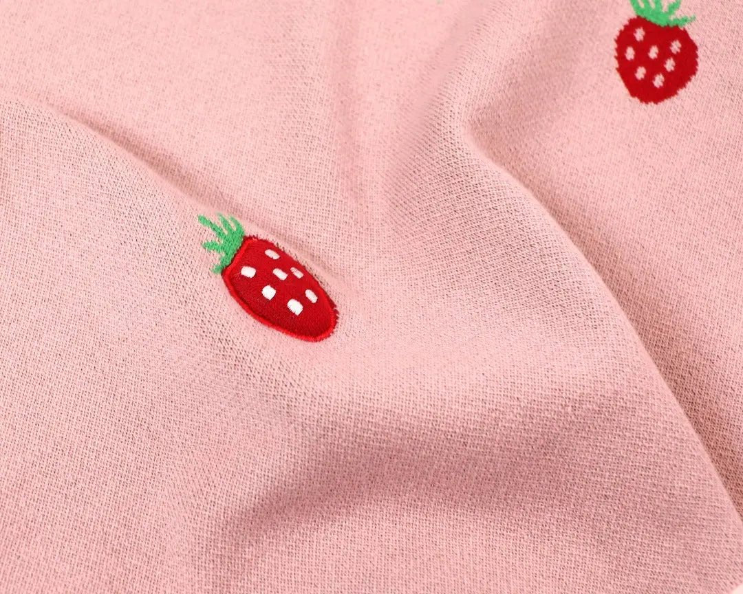 Close-up of a soft pink knitted baby blanket featuring small red strawberry motifs with green leaves and white seed details. The jacquard design adds a playful and textured look to the fabric, which appears smooth and cozy. The charming pattern gives the blanket a sweet, whimsical feel, perfect for a nursery or baby gift