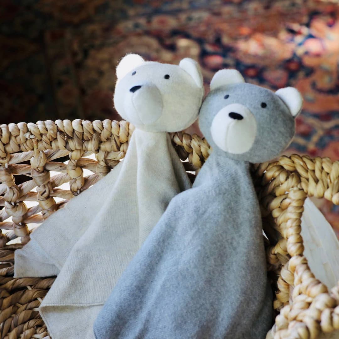 Two soft bear lovey security blankets, one cream and one gray, sit side by side in a woven basket with a warm, cozy background.