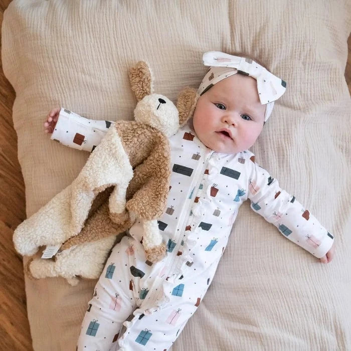 Baby lying on a beige blanket, dressed in a white onesie with gift box prints and a matching headband, cuddling a plush dog lovey.