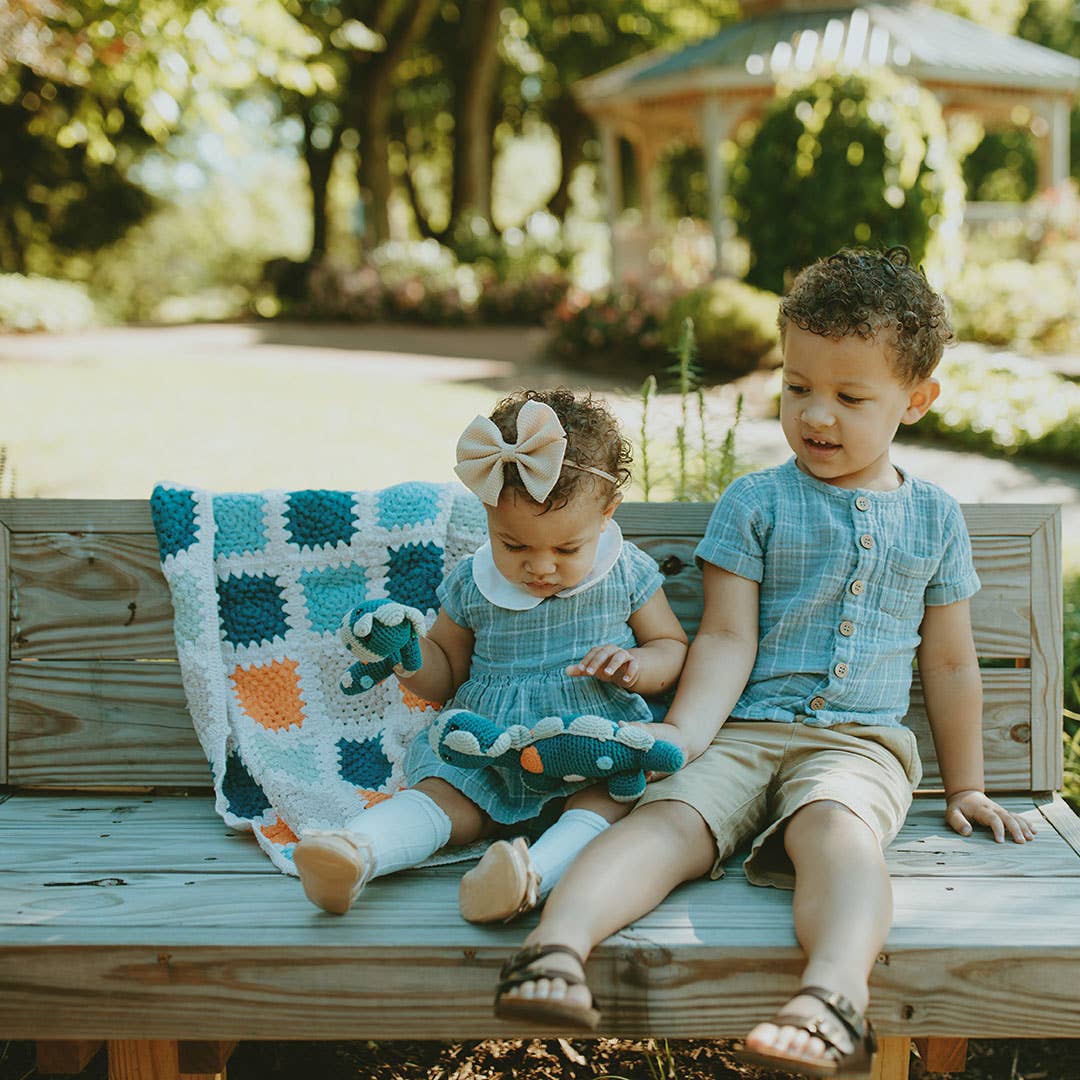 Two young children sitting on a wooden bench in a lush garden, with a hand-crocheted blanket featuring blue and orange squares next to them. The girl, wearing a light blue dress and a matching bow, is holding a knitted toy, while the boy, dressed in a buttoned blue shirt and shorts, looks at her.