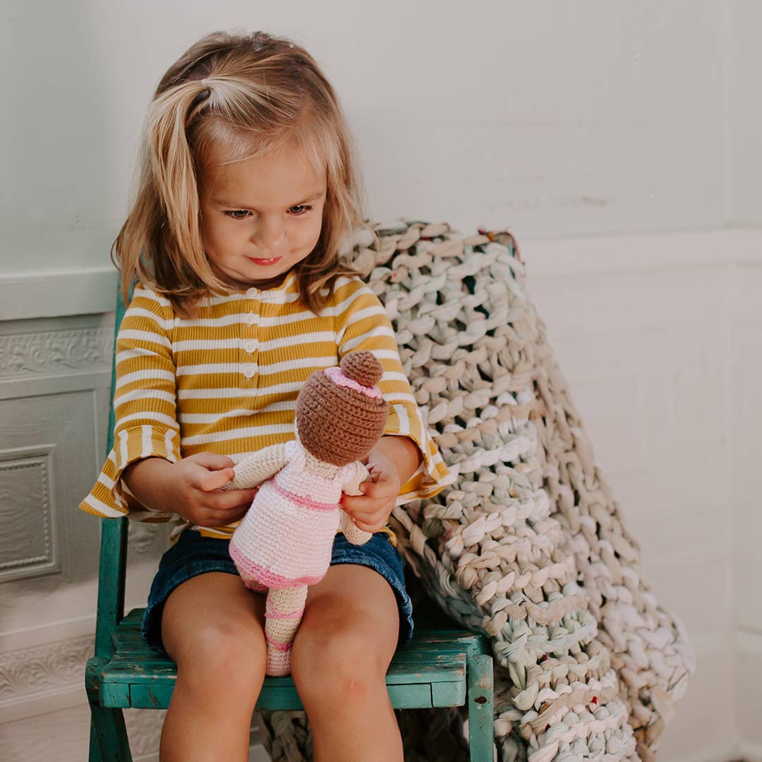 This image shows a young girl sitting on a teal wooden chair, wearing a yellow and white striped top with denim shorts. She is holding a small crocheted doll dressed in pink, with a brown bun on its head, while gazing at it intently. A chunky knitted blanket is draped over the chair, adding a cozy, homey feel to the scene. The child’s expression reflects concentration and affection towards the doll.