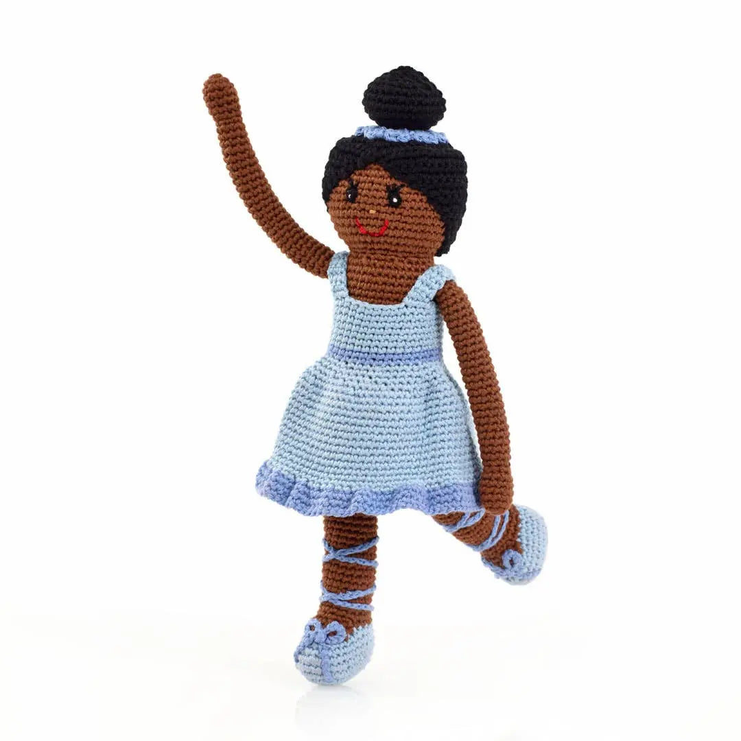 This image features a handmade crocheted ballerina doll dressed in a light blue dress and matching ballet slippers. The doll has brown skin, with her black hair styled in a bun and adorned with a matching blue headband. Crafted from organic cotton, the doll stands in a playful, dancing pose, with one arm raised and one leg lifted, showcasing its charming and whimsical design.