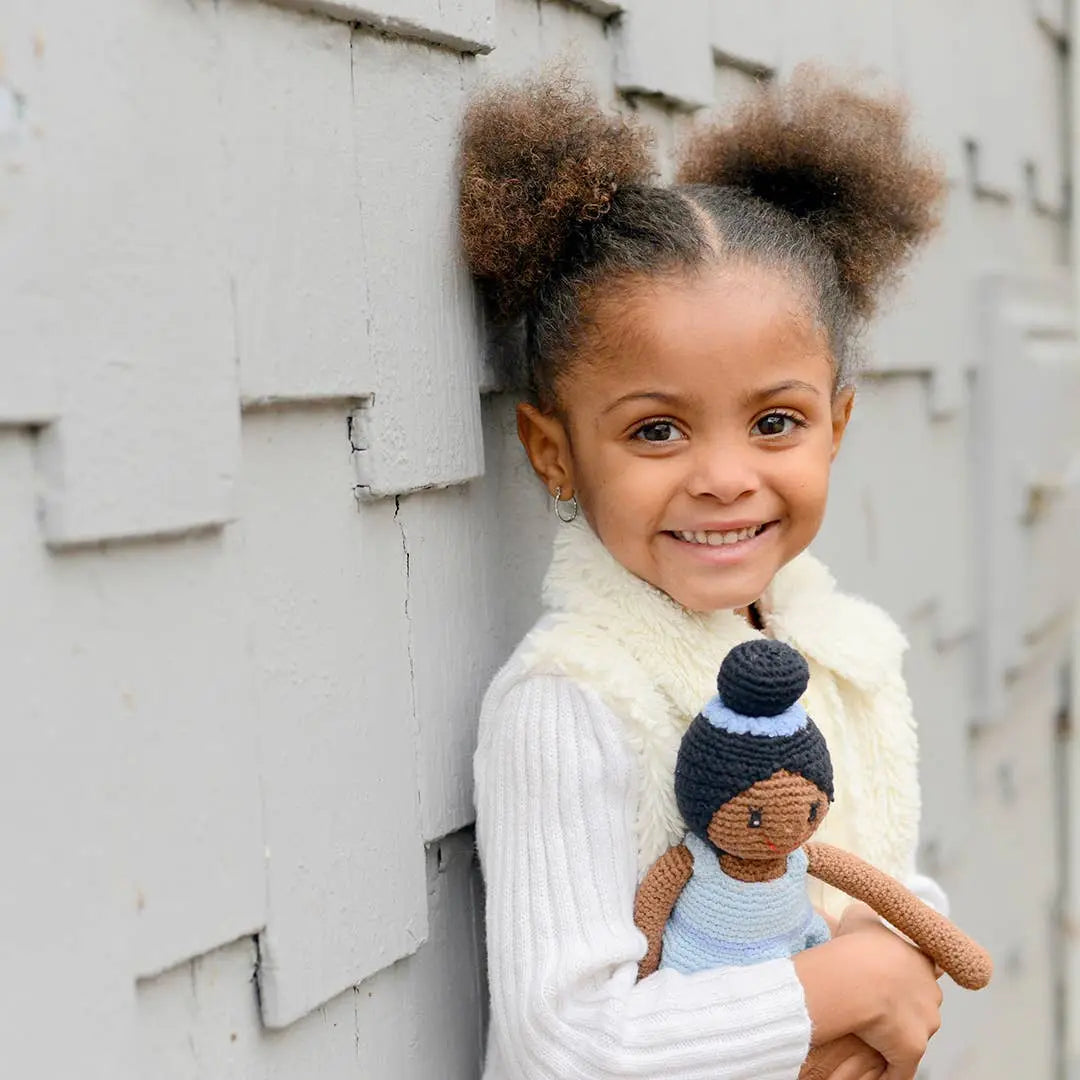 This image shows a young girl smiling while holding a crocheted ballerina doll dressed in blue. The girl, wearing a cream-colored vest and a white sweater, has her hair styled in two puff ponytails. She leans against a textured, gray wooden wall, radiating joy and warmth as she clutches the handmade doll. The scene captures a heartwarming moment of playfulness and comfort.