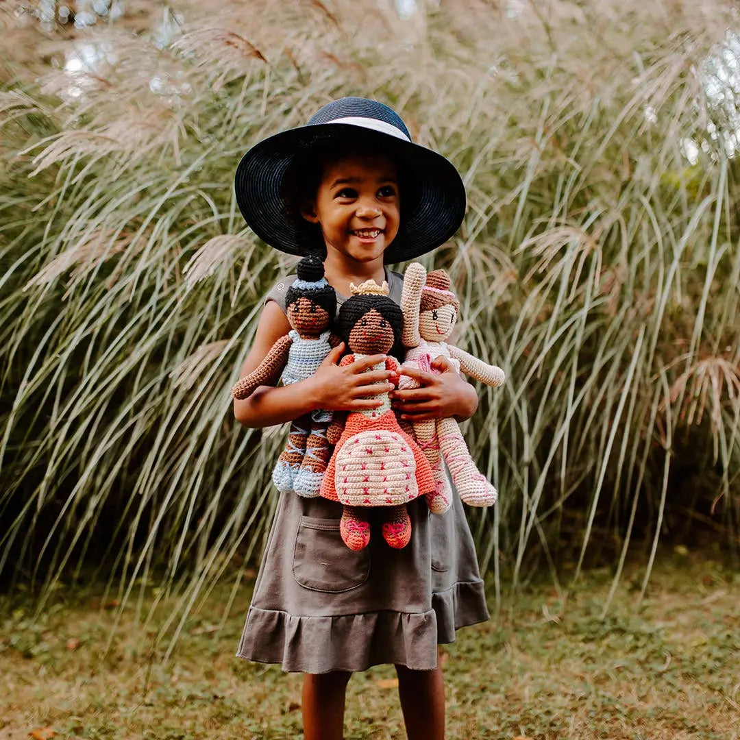 This image captures a young girl standing outdoors, joyfully holding several handmade crocheted dolls in her arms. She wears a wide-brimmed black hat and a simple gray dress with pockets. The dolls are dressed in various colors, with one ballerina in blue among them. Behind her, tall grasses sway, adding a natural, earthy backdrop to the playful and whimsical scene. The girl's wide smile radiates happiness and warmth.