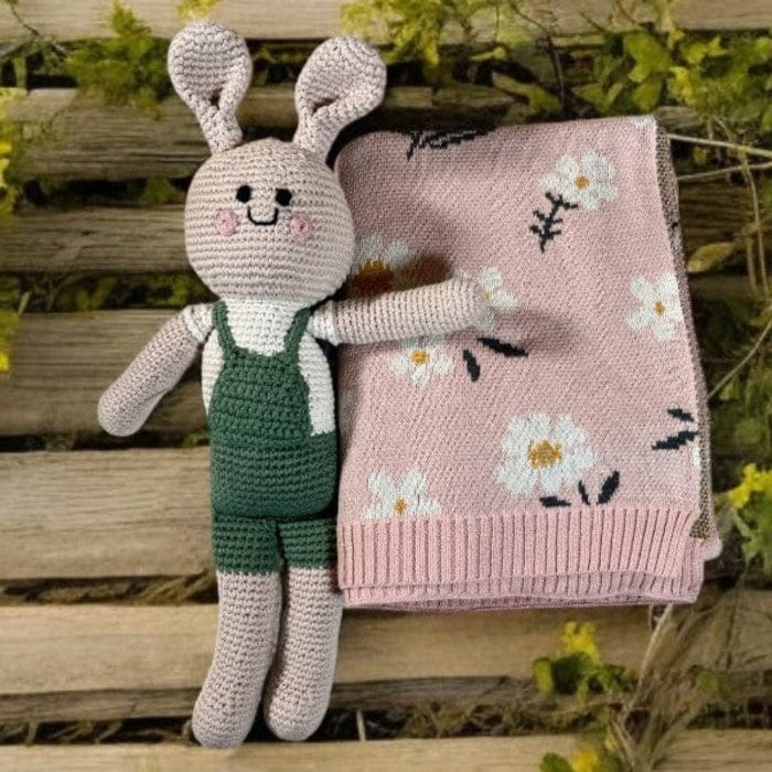 A crocheted bunny toy in green overalls lies next to a folded pink blanket with white daisy flower patterns on a wooden surface.
