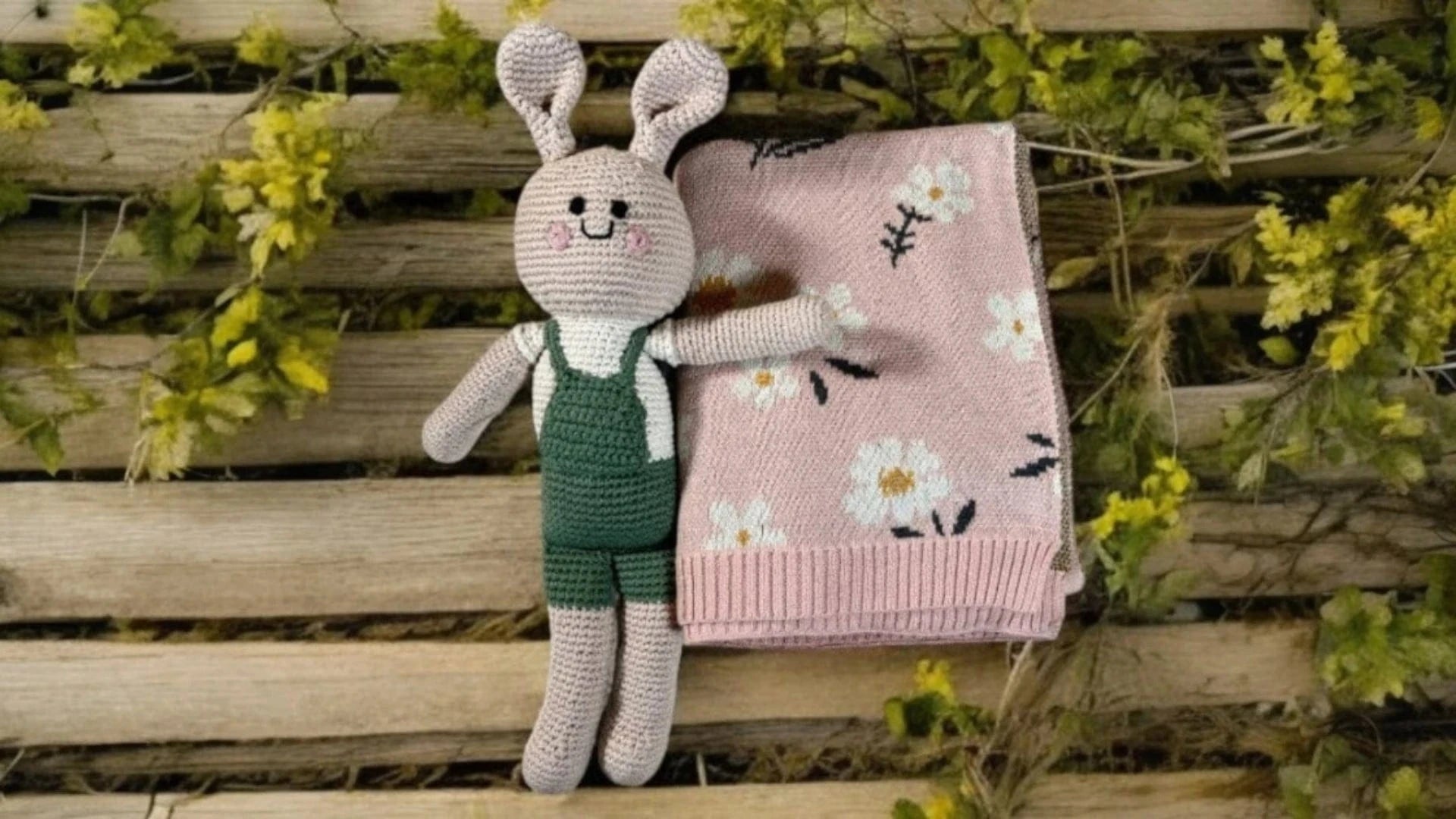 Knitted bunny toy with a pink blanket featuring floral patterns on a wooden surface with greenery.