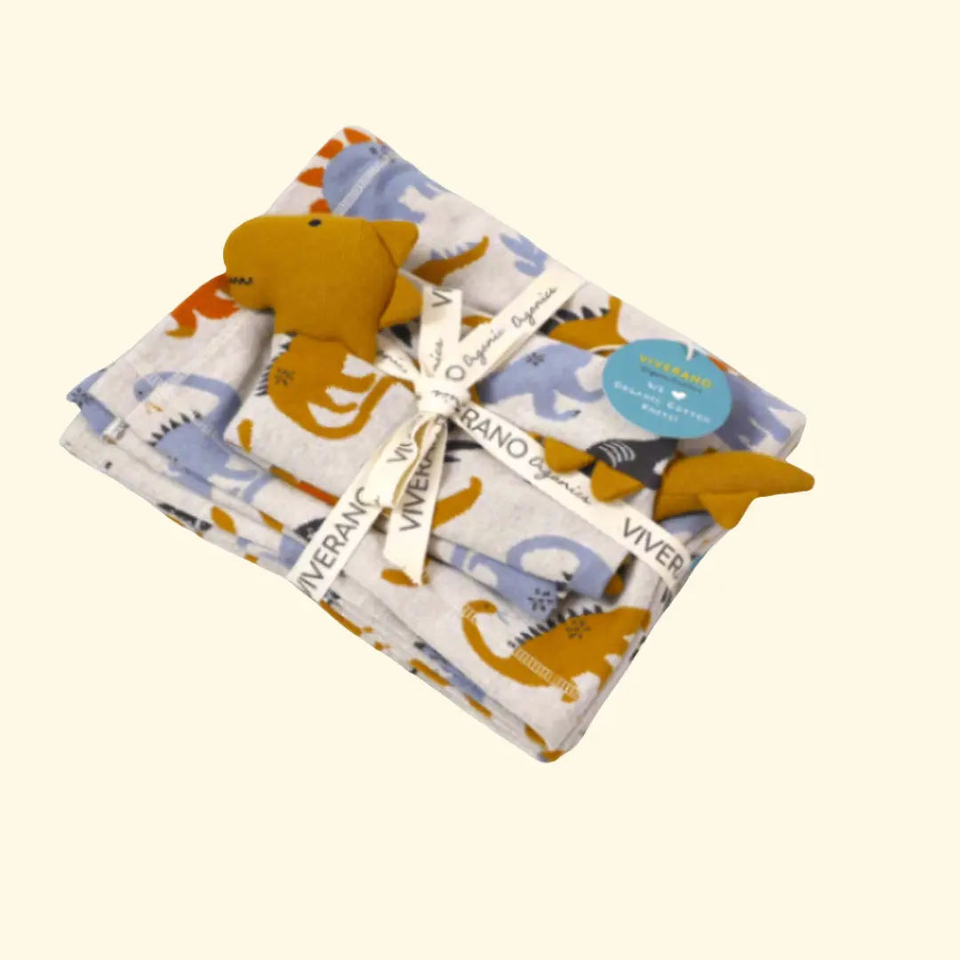 A set of folded baby blankets with dinosaur prints is neatly tied with a ribbon labeled "Viverano Organic". The blankets are accompanied by a small dinosaur plush toy. Pink, blue, yellow, and purple balloons are scattered around, adding a festive touch to the scene.