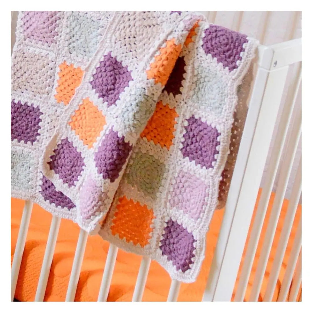 A hand-crocheted baby blanket in purple, orange, and white squares draped over the side of a white crib, adding a cozy touch to the nursery.