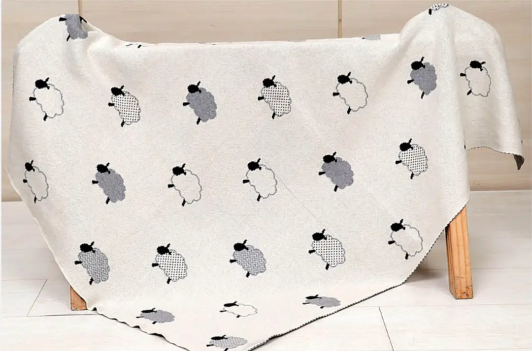 This image features a baby blanket draped over a wooden surface, showcasing a delightful sheep pattern. The blanket, made from organic cotton, displays black, white, and gray sheep scattered across a soft beige background. The whimsical design adds a cozy and playful touch, making it perfect for a baby’s nursery or as a snuggly accessory. The use of neutral tones makes it a versatile piece for various nursery themes.