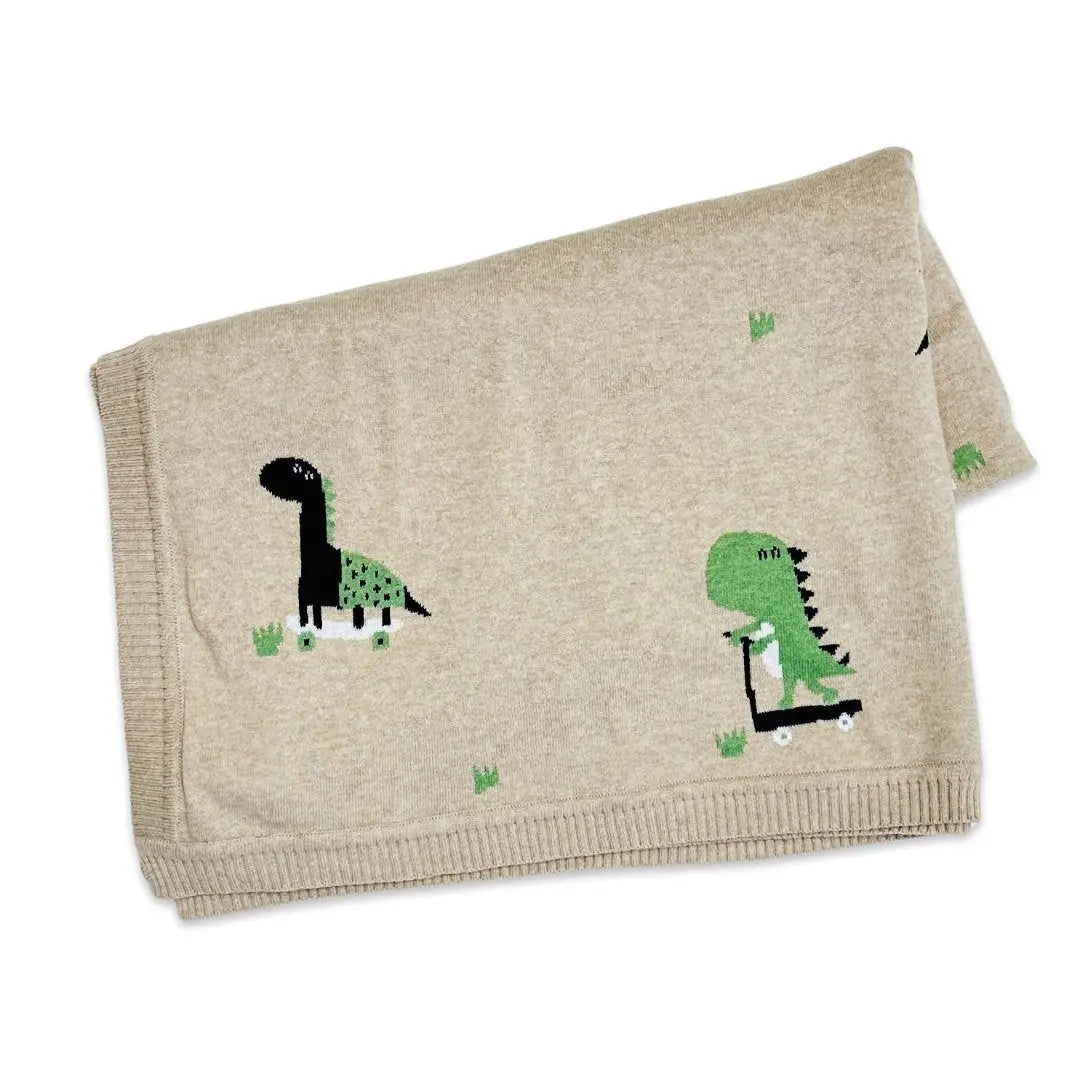 This image shows a folded beige baby blanket made from organic cotton, featuring a cute dinosaur-themed design. One dinosaur is shown riding a skateboard, while another is on a scooter, both in green and black with small grass details scattered across the blanket. The playful, modern design makes this blanket perfect for adding a fun touch to a nursery or for any baby who loves dinosaurs. The ribbed edges provide a neat and cozy finish to the blanket.