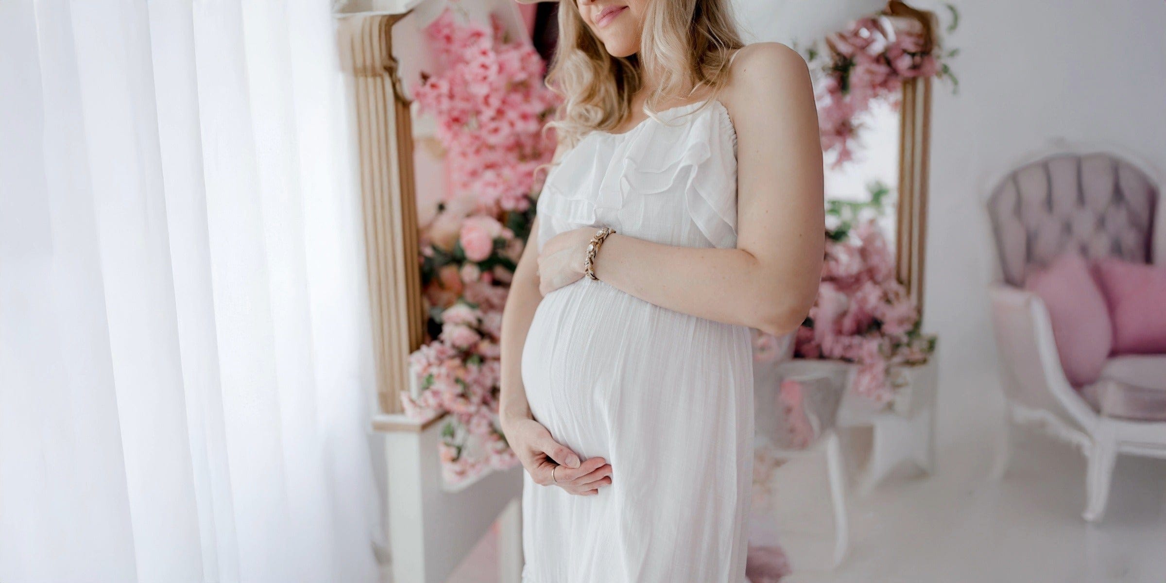 Pregnant woman in a white dress standing in a decorated room with pink flowers and a mirror.