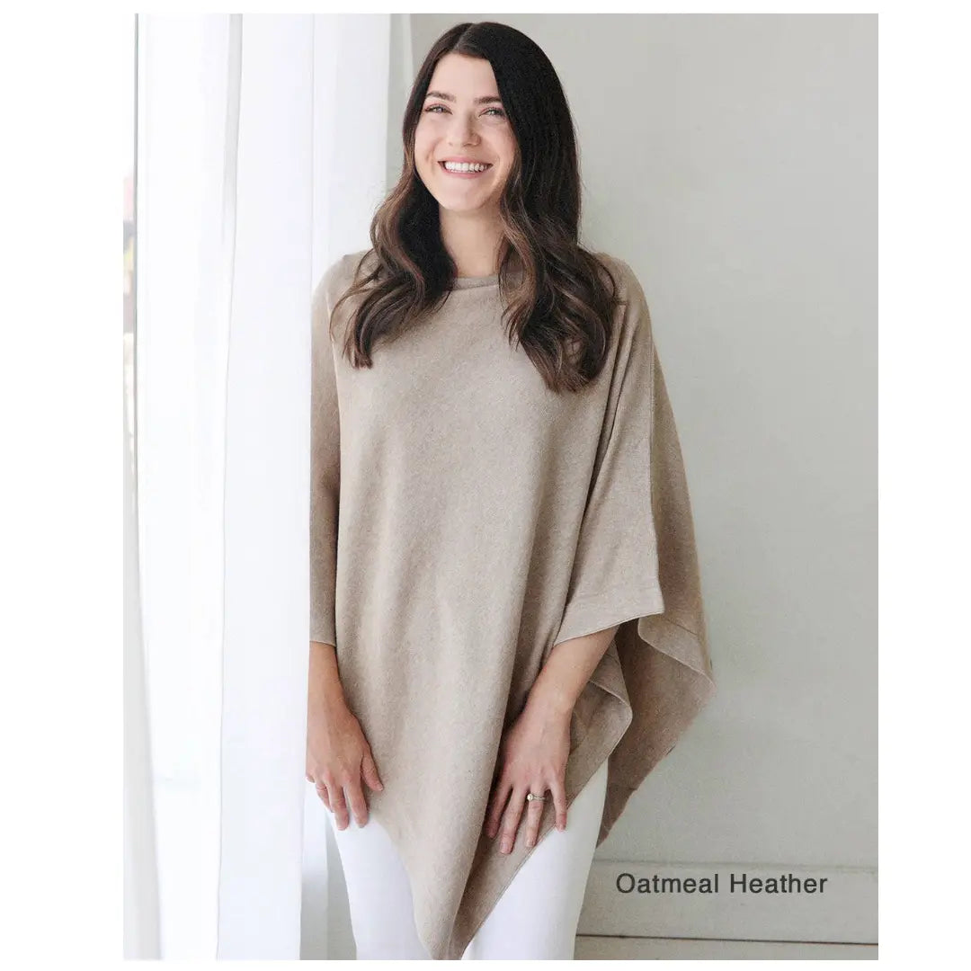 This image shows a woman standing in a well-lit room, smiling while wearing a beige-colored poncho labeled "Oatmeal Heather" in the corner. The garment is flowy and relaxed, pairing with white pants, giving a casual and comfortable look.