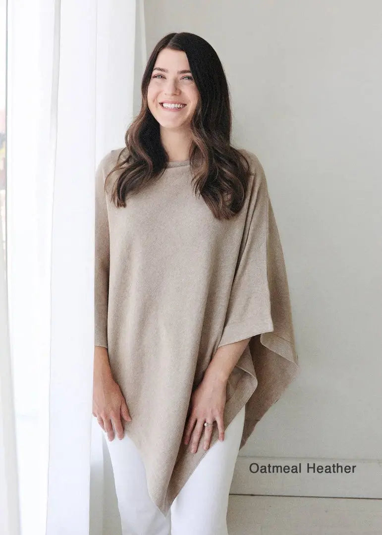 This image depicts a woman standing near a window with soft, natural light streaming in. She is wearing a beige poncho over black pants, looking contemplatively to the side. The simple, cozy outfit and serene lighting give the image a calm and minimalist feel. A plant in the corner adds a subtle, natural element to the setting.