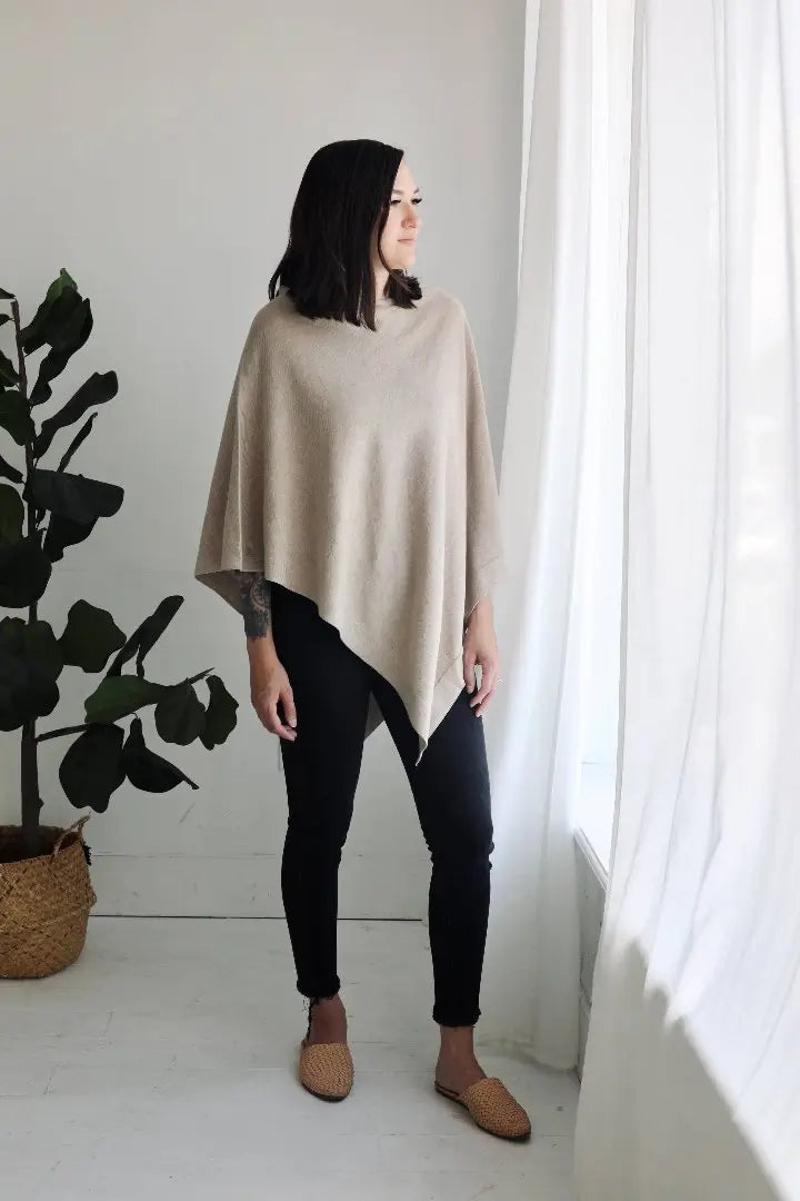 This image shows a woman standing near a window in a minimalistic room, wearing a light beige poncho over black skinny pants. She pairs the outfit with woven, tan-colored flats, giving off a cozy, casual vibe. A large green plant in a woven basket adds a natural element to the scene.