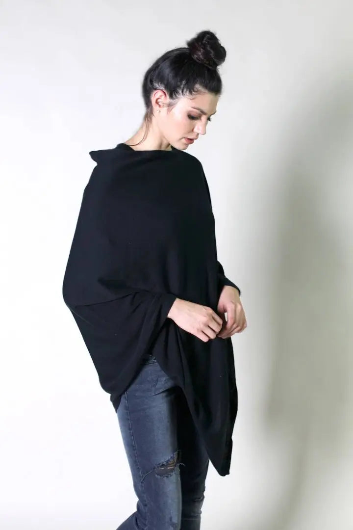 This image features a woman wearing a black poncho with a loose, relaxed fit. She pairs it with dark, ripped jeans for a casual and understated look. Her hair is styled in a high bun, and her gaze is directed downward, contributing to a minimalist and effortless aesthetic.