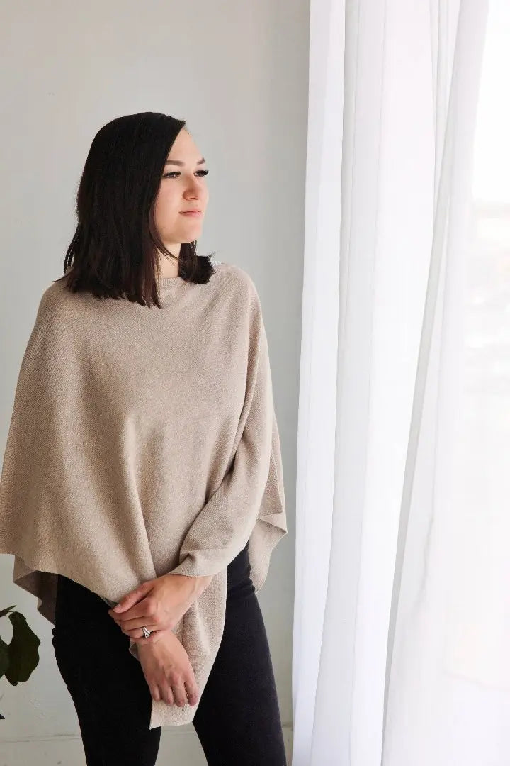 This image depicts a woman standing near a window with soft, natural light streaming in. She is wearing a beige poncho over black pants, looking contemplatively to the side. The simple, cozy outfit and serene lighting give the image a calm and minimalist feel. A plant in the corner adds a subtle, natural element to the setting.