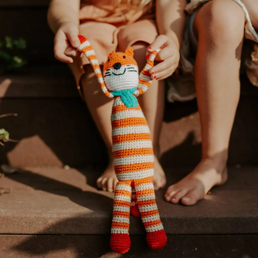 This image captures a playful moment where a child holds a handmade crocheted fox toy by its arms. The fox, made from organic cotton, is striped in orange and white, with red feet and a turquoise scarf around its neck. It has a cheerful, smiling face and serves as a charming woodland-themed toy. The setting appears to be outdoors, as the child sits on steps with bare feet, adding to the relaxed and joyful atmosphere of playtime.