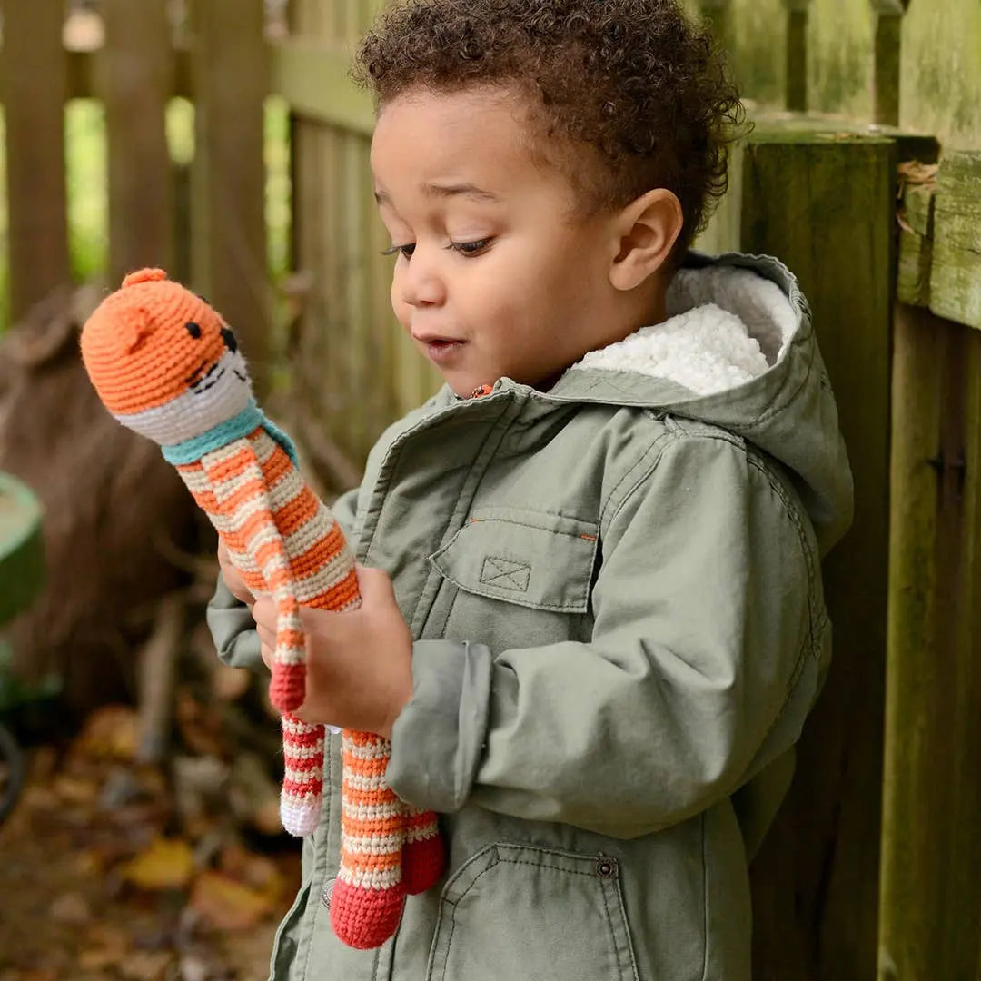 This image shows a young child outdoors, holding a handmade crocheted fox toy. The fox is orange and white with red feet and a turquoise scarf, crafted from organic cotton. The child, dressed warmly in a green jacket with a cozy fleece lining, appears to be examining the toy with curiosity and delight. The wooden fence and autumn leaves in the background create a natural, playful setting.