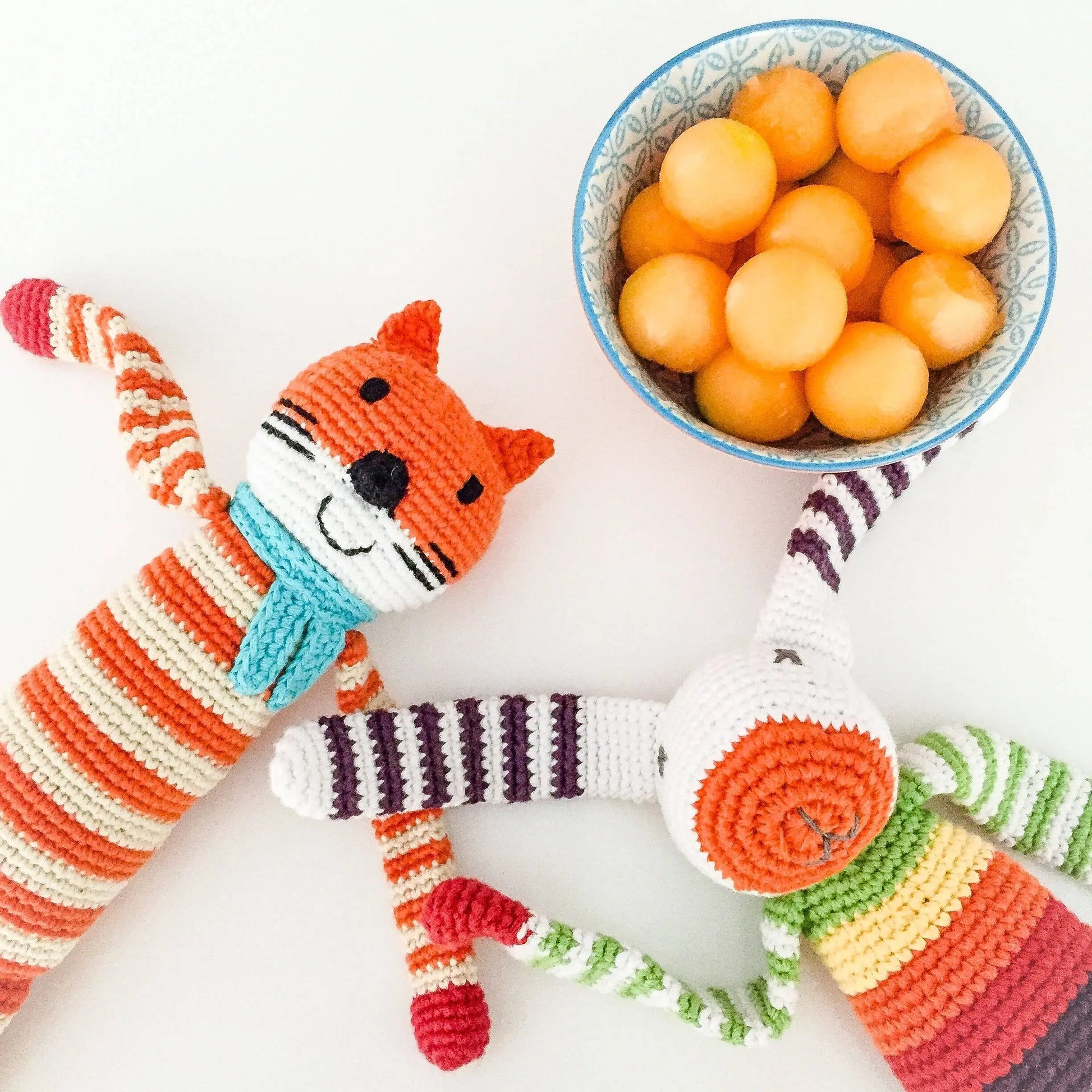 This image features two handmade crocheted toys placed next to a bowl of melon balls. One toy is a striped orange and white fox with a turquoise scarf, and the other is a whimsical figure with rainbow stripes, long ears, and a playful design. Both toys are made from organic cotton and are designed to be soft and child-friendly. The bright colors of the toys and the fruit add a fun and vibrant touch to the scene, creating a cheerful and playful mood.