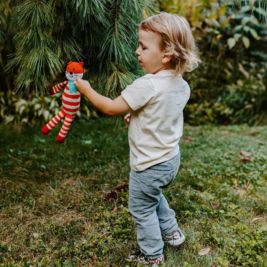 This image features a young child standing in a grassy outdoor setting, holding a handmade crocheted fox toy. The fox is striped in red and white, with a turquoise scarf, crafted from organic cotton. The child appears to be gently interacting with a low-hanging pine branch, exploring the natural surroundings. The scene captures a peaceful and curious moment of outdoor play, with the lush greenery adding to the serene, nature-inspired atmosphere.