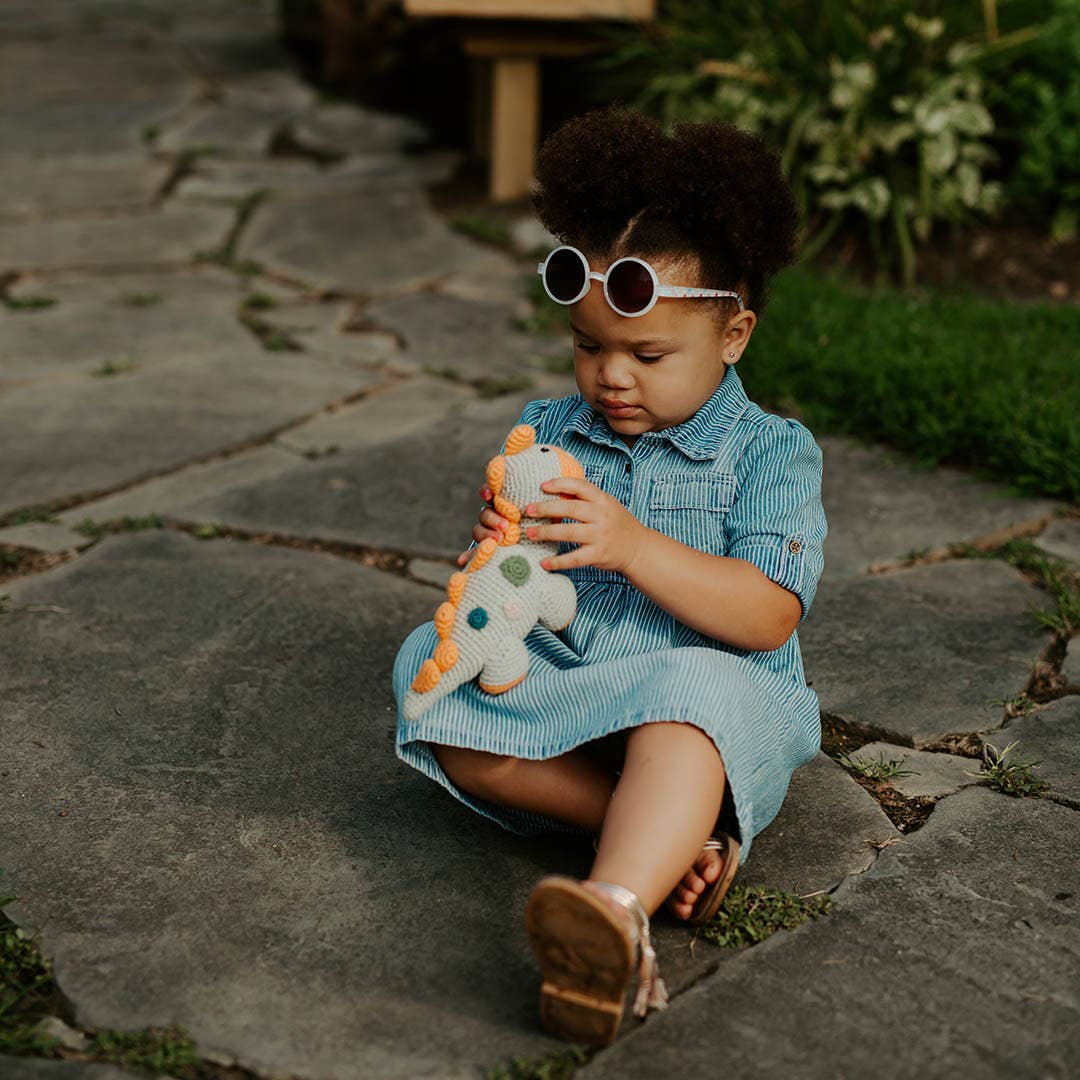 Child in a blue dress sitting on stone steps holding a dinosaur toy, wearing sunglasses.