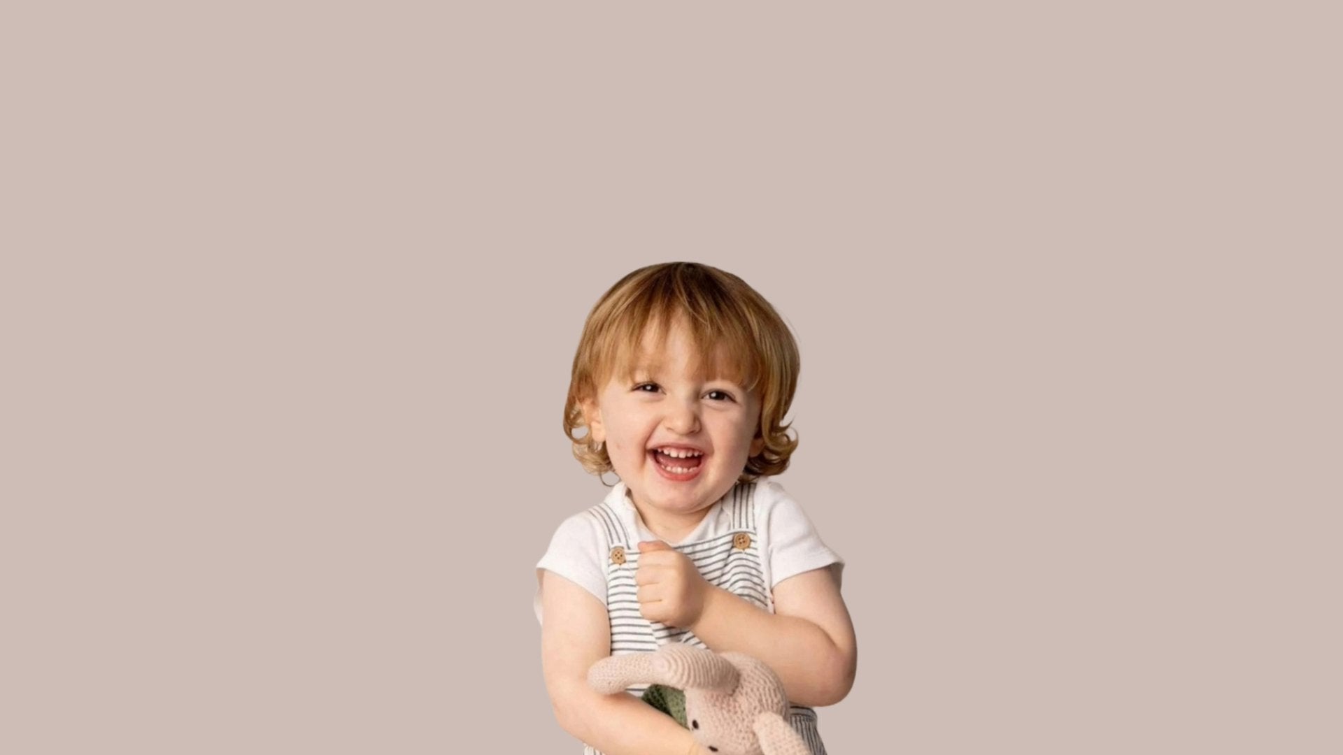 A smiling toddler with light brown hair wears striped overalls over a white shirt and holds a knitted stuffed animal while laughing. 